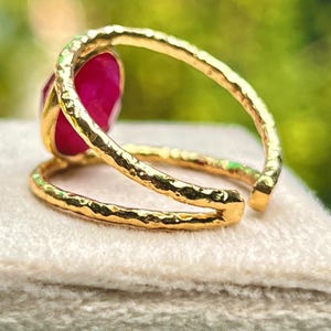 K&ouml;nnte beinhalten: Ein goldfarbener Ring mit einem gro&szlig;en, oval geformten, tiefrosa Edelstein. Der Ring hat ein strukturiertes Band und ein Doppelband-Design. Der Edelstein ist in einer goldenen L&uuml;nette gefasst, pr&auml;sentiert auf einer hellen Oberfl&auml;che.