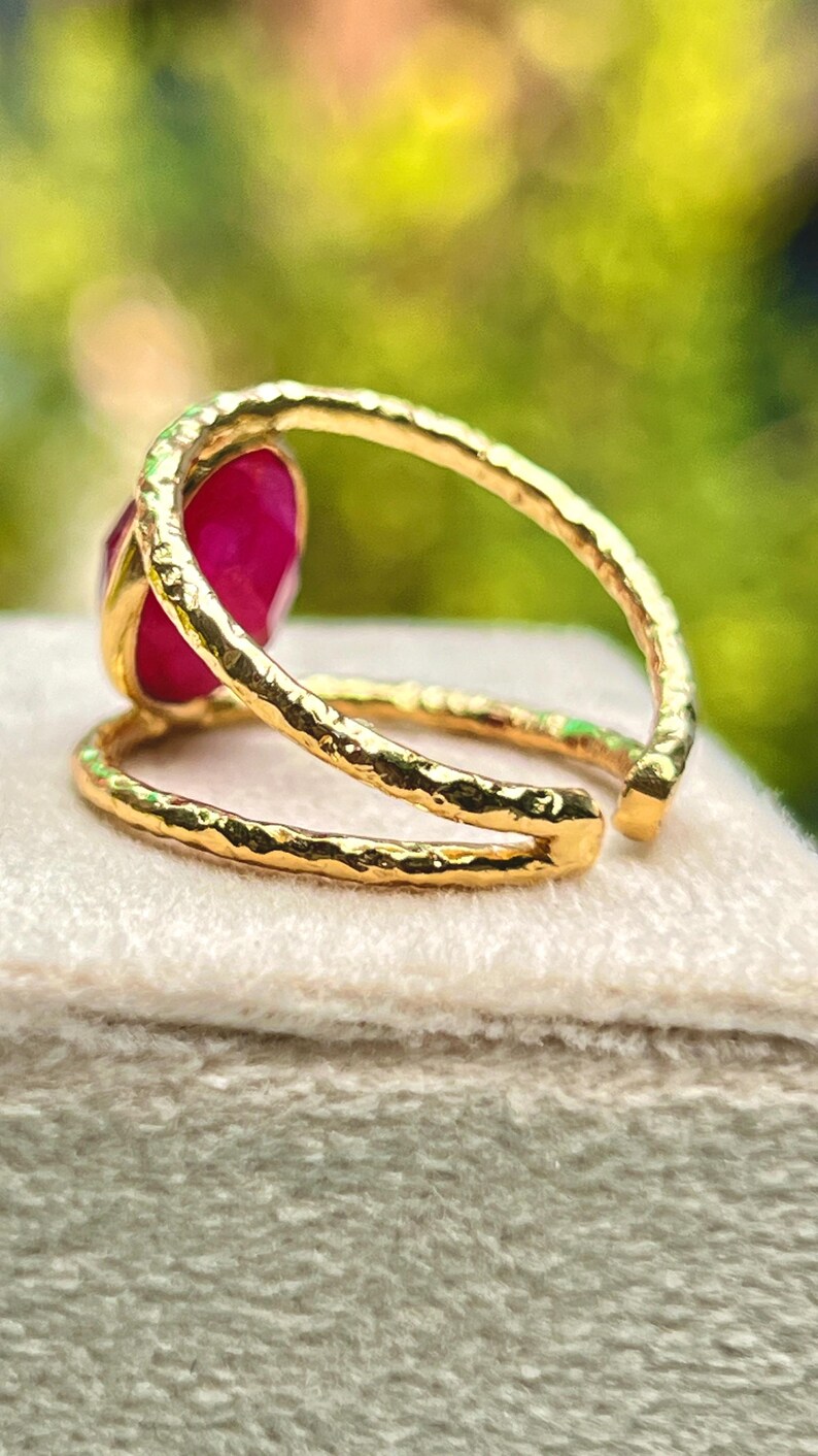 K&ouml;nnte beinhalten: Ein goldfarbener Ring mit einem gro&szlig;en, oval geformten, tiefrosa Edelstein. Der Ring hat ein strukturiertes Band und ein Doppelband-Design. Der Edelstein ist in einer goldenen L&uuml;nette gefasst, pr&auml;sentiert auf einer hellen Oberfl&auml;che.