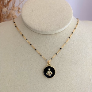 Può includere: Una collana con catena dorata con un ciondolo in smalto nero con un design di ape dorata.