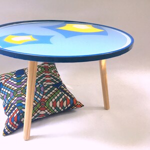 May include: A round blue table with a yellow and blue abstract design on the top. The table has four wooden legs and is sitting on a white surface.