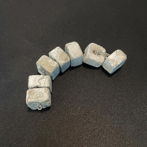 May include: A bracelet composed of six rectangular, light grey concrete blocks with a rough, porous texture. The blocks are connected by a thin, silver-toned metal chain that runs through the centre of each piece, creating a flexible, curved link design.