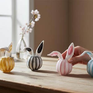 May include: Four decorative ceramic bunny figurines with striped bodies and upright ears. The bunnies are in yellow, black and white, pink, and blue. The ears have gold, black, pink, and blue accents. They are arranged on a wooden table.