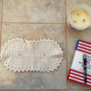 Könnte beinhalten: Ein cremefarbenes, oval gehäkeltes Deckchen mit einem gewellten Rand. Eine brennende Kerze in einem floral bedruckten Glas und ein Buch mit dem Titel "American History in Bite-Sized Chunks" mit einem Stift sind ebenfalls vorhanden.