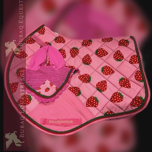 May include: A pink quilted horse saddle pad and matching ear bonnet set featuring a repeating strawberry pattern. The saddle pad has a green braided trim and a rectangular patch with the text BURAQ EQUESTRIANS embroidered in gold thread.
