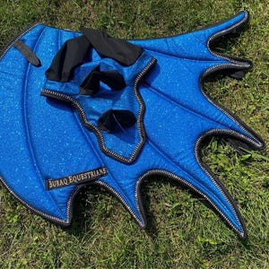 May include: A bright blue glittery horse fly mask with attached wing-shaped ear covers. The fabric features a sparkling finish with black trim and black ear sections. A black label with the white text BURAQ EQUESTRIANS is stitched onto the side.