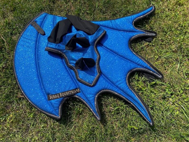 May include: A bright blue glittery horse fly mask with attached wing-shaped ear covers. The fabric features a sparkling finish with black trim and black ear sections. A black label with the white text BURAQ EQUESTRIANS is stitched onto the side.