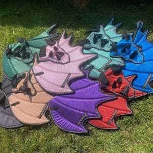 May include: A collection of glittery, wing-shaped horse ear bonnets in various colours including green, pink, blue, red, purple, and black. Each bonnet features a structured, bat-like wing design with black trim and a small black label reading JURAQ EQUESTRIAN.