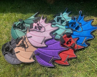 May include: A collection of glittery, wing-shaped horse ear bonnets in various colors including green, pink, blue, red, purple, and black. Each bonnet features a structured, bat-like wing design with black trim and a small black label reading JURAQ EQUESTRIAN.