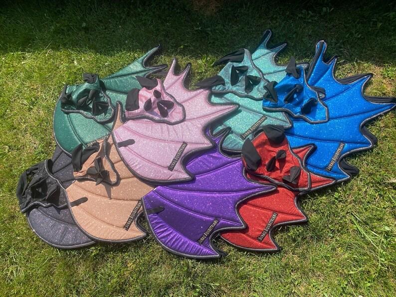 May include: A collection of glittery, wing-shaped horse ear bonnets in various colors including green, pink, blue, red, purple, and black. Each bonnet features a structured, bat-like wing design with black trim and a small black label reading JURAQ EQUESTRIAN.
