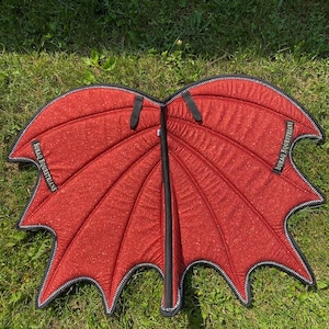May include: A pair of red glittery fabric wings with a black trim and a central black spine. The wings feature a segmented, bat-like shape with pointed edges. A black fabric label with the white text BURAQ EQUESTRIANS is stitched onto each wing.