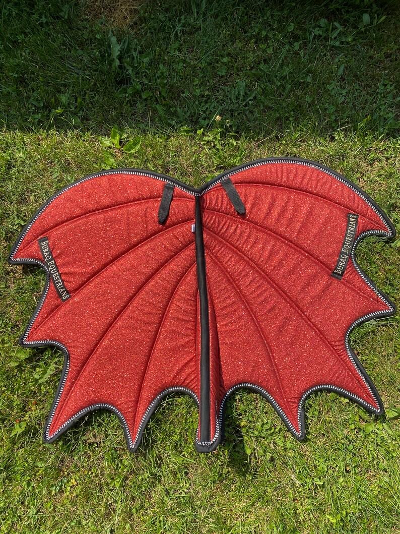 May include: A pair of red glittery fabric wings with a black trim and a central black spine. The wings feature a segmented, bat-like shape with pointed edges. A black fabric label with the white text BURAQ EQUESTRIANS is stitched onto each wing.