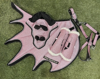 May include: A pink equestrian set with glitter accents, including a saddle pad, leg wraps, and a halter. The saddle pad has a unique wing-like shape with black trim. The set is designed for horses and features the text "BURAQ EQUESTRIANS".