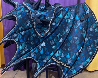 May include: A blue and teal dragon-themed horse saddle pad featuring a scale pattern, black trim, and pointed wing-like extensions. The text BURAQ EQUESTRIANS is printed in white on the lower section of the pad, which is mounted on a black stand.
