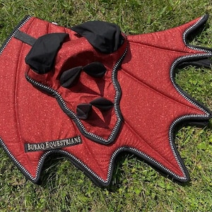 May include: A red glittery equestrian saddle pad with black trim and black fabric accents. The pad features a distinct wing-like shape with pointed edges and a black label with the white text BURAQ EQUESTRIANS stitched onto the lower left corner.
