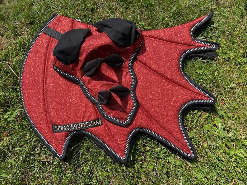 May include: A red glittery equestrian saddle pad with black trim and black fabric accents. The pad features a distinct wing-like shape with pointed edges and a black label with the white text BURAQ EQUESTRIANS stitched onto the lower left corner.