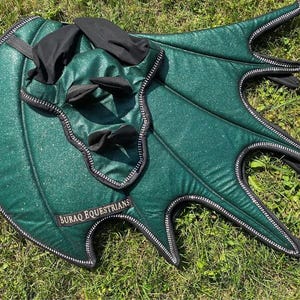 May include: A dark green glittery horse ear bonnet and matching saddle pad set featuring black trim and black fabric accents. The saddle pad has a wing-like shape with multiple points and a black label that reads BURAQ EQUESTRIANS in gold text.