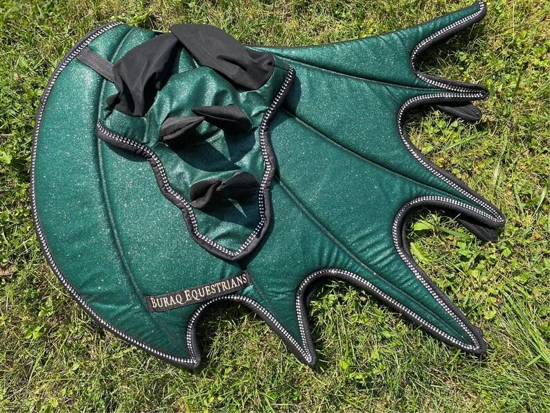 May include: A dark green glittery horse ear bonnet and matching saddle pad set featuring black trim and black fabric accents. The saddle pad has a wing-like shape with multiple points and a black label that reads BURAQ EQUESTRIANS in gold text.