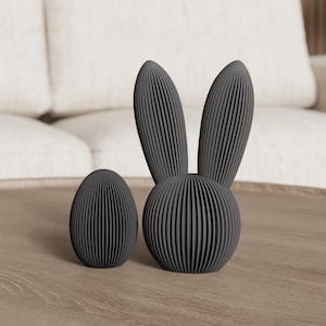 Può includere: Un set decorativo grigio scuro a forma di coniglio e uovo. Il coniglio ha lunghe orecchie e un corpo rotondo. Sia il coniglio che l'uovo hanno un design a coste. Gli oggetti sono esposti su un tavolo di legno.