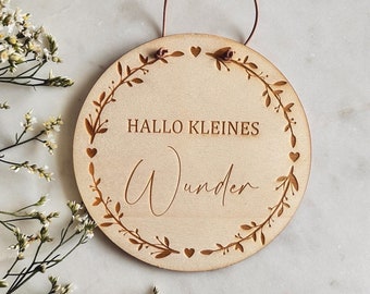 Peut inclure: Panneau rond en bois clair avec l'inscription gravée "HALLO KLEINES Wunder", entourée d'une couronne florale. Il est orné de cœurs et de petites fleurs. Une ficelle marron permet de l'accrocher.