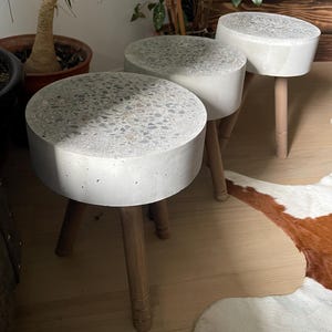 May include: Three small, round stools with concrete tops and wooden legs. The concrete tops are light gray with a speckled pattern. The wooden legs are light brown and slightly tapered. The stools are arranged in a row, with the tops facing the viewer.