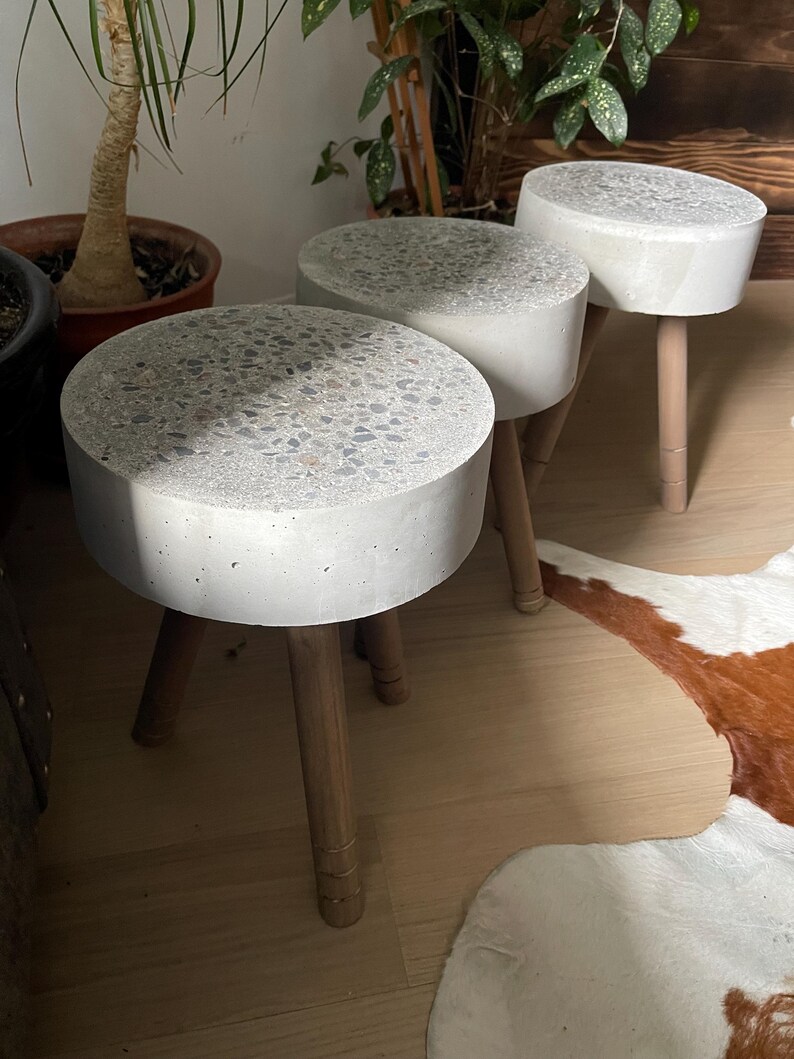 May include: Three small, round stools with concrete tops and wooden legs. The concrete tops are light gray with a speckled pattern. The wooden legs are light brown and slightly tapered. The stools are arranged in a row, with the tops facing the viewer.