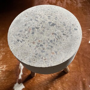 May include: A round, gray concrete side table with a speckled surface. The table has a cylindrical base and three wooden legs. The table is shown on a brown and white patterned surface.