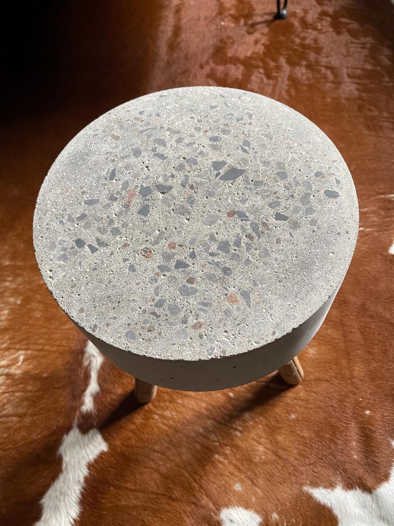 May include: A round, gray concrete side table with a speckled surface. The table has a cylindrical base and three wooden legs. The table is shown on a brown and white patterned surface.