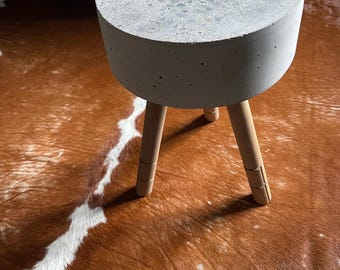 May include: A small, round side table with a concrete top and three wooden legs. The tabletop is speckled with various colors. The legs are light brown and angled outward for stability. The table is a modern design.