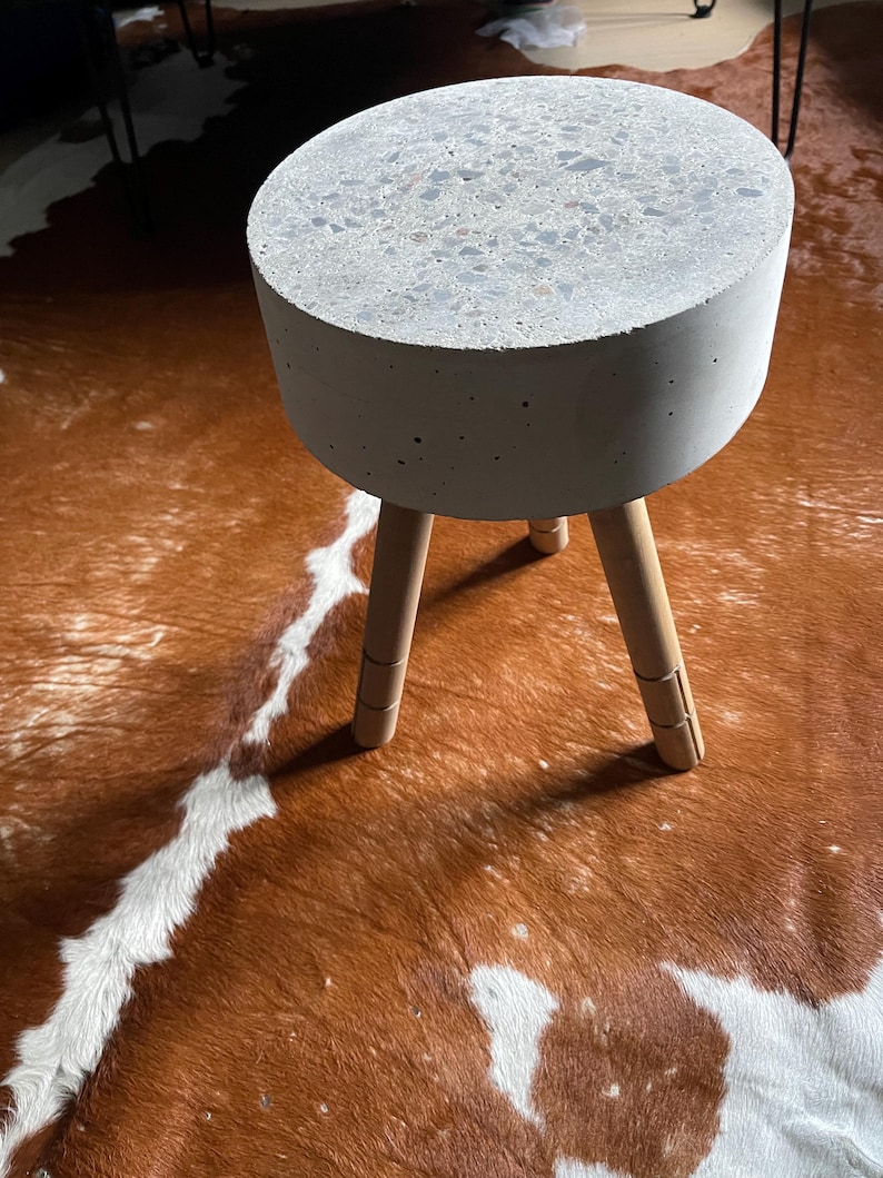 May include: A small, round side table with a concrete top and three wooden legs. The tabletop is speckled with various colors. The legs are light brown and angled outward for stability. The table is a modern design.