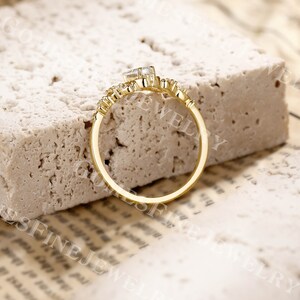 Op de afbeelding: Een goudkleurige ring met een rechthoekige, heldere edelsteen in het midden. De ring heeft een delicaat ontwerp met kleinere, ronde edelstenen. De ring is geplaatst op een getextureerd, lichtgekleurd oppervlak.
