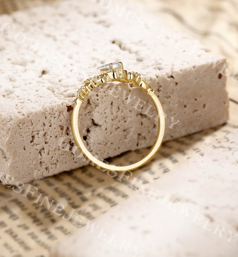 Op de afbeelding: Een goudkleurige ring met een rechthoekige, heldere edelsteen in het midden. De ring heeft een delicaat ontwerp met kleinere, ronde edelstenen. De ring is geplaatst op een getextureerd, lichtgekleurd oppervlak.
