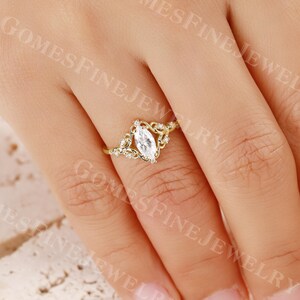 Op de afbeelding: Een gouden ring met een heldere, marquise-geslepen edelsteen. De ring heeft een delicaat, filigraan ontwerp met kleine, ronde accentstenen. De ring is geplaatst tegen een neutrale achtergrond.