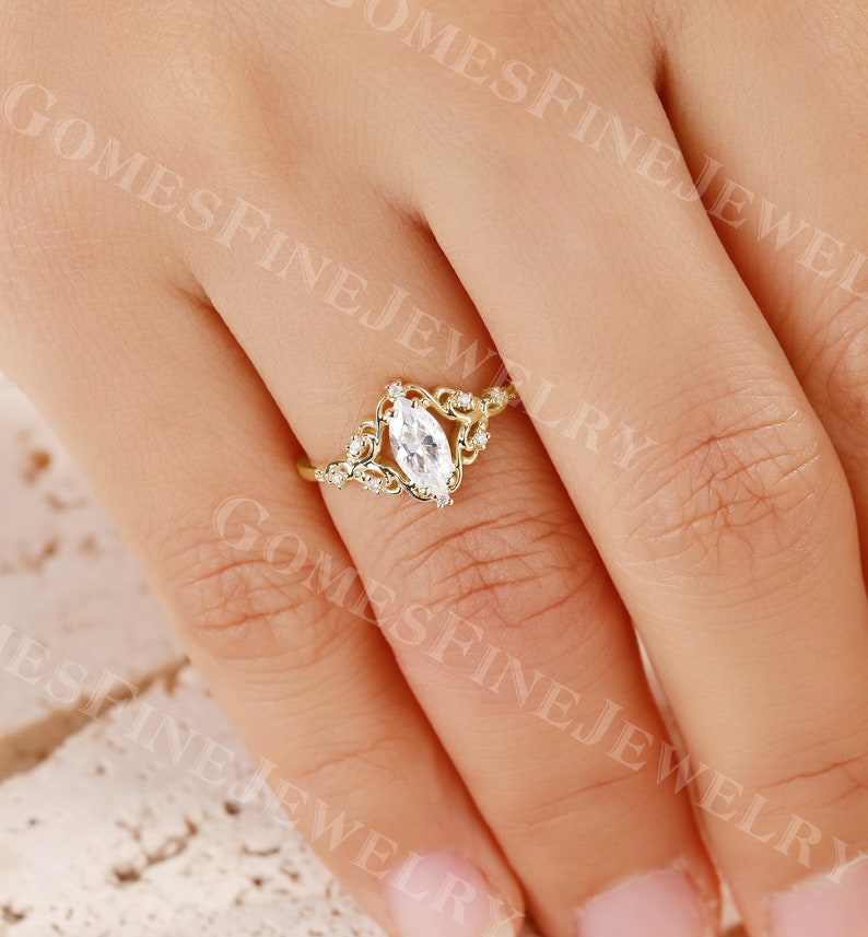Op de afbeelding: Een gouden ring met een heldere, marquise-geslepen edelsteen. De ring heeft een delicaat, filigraan ontwerp met kleine, ronde accentstenen. De ring is geplaatst tegen een neutrale achtergrond.