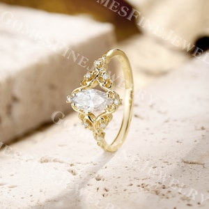 Op de afbeelding: Een goudkleurige ring met een heldere, marquise-geslepen edelsteen als middelpunt. De ring heeft ingewikkelde filigraan details rond de edelsteen en kleinere accentstenen. De band is eenvoudig, gepolijst goud. De ring staat tegen een neutrale, getextureerde achtergrond.