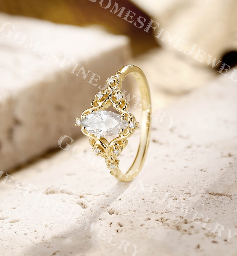 Op de afbeelding: Een goudkleurige ring met een heldere, marquise-geslepen edelsteen als middelpunt. De ring heeft ingewikkelde filigraan details rond de edelsteen en kleinere accentstenen. De band is eenvoudig, gepolijst goud. De ring staat tegen een neutrale, getextureerde achtergrond.