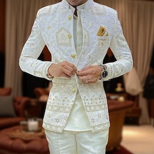 Può includere: Un completo bianco ricamato con dettagli dorati. La giacca presenta intricati motivi floreali e geometrici, bottoni dorati e un fazzoletto da taschino. Il completo include pantaloni abbinati e una camicia con colletto. L'orologio al polso ha un quadrante nero e un cinturino argentato.