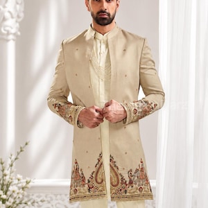 Op de afbeelding: Een beige sherwani-jas met ingewikkeld borduurwerk op de manchetten en zoom, gecombineerd met een bijpassend lichtcrèmekleurig overhemd en broek. De jas toont gedetailleerde patronen van figuren en paisley-motieven in rode, gouden en groene tinten.