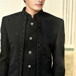 May include: A black formal jacket with a mandarin collar and decorative embellishments. The jacket features a series of silver buttons down the front and intricate detailing along the edges and down the front. The fabric has a subtle sheen, adding to its formal appearance.