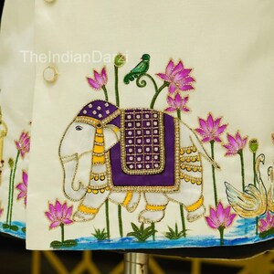 May include: Ivory-colored embroidered jacket featuring an elephant with a purple saddle, a green bird, and pink lotus flowers. The jacket has gold-colored buttons and trim. The design includes a water scene with swans.
