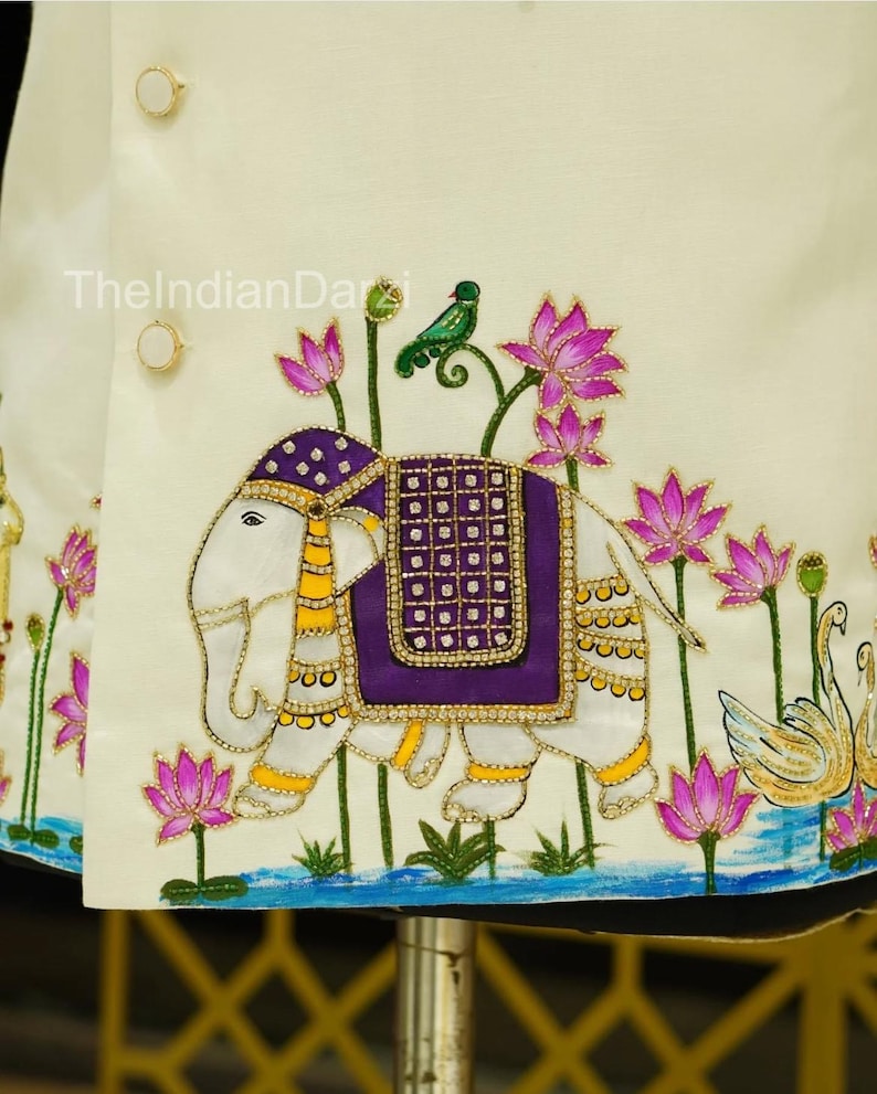 May include: Ivory-colored embroidered jacket featuring an elephant with a purple saddle, a green bird, and pink lotus flowers. The jacket has gold-colored buttons and trim. The design includes a water scene with swans.