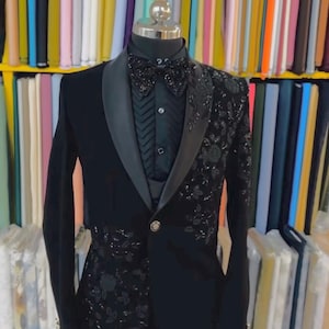 May include: A black tuxedo with a satin lapel and a patterned design on the right side. The tuxedo includes a black shirt, bow tie, and a single-button closure. The background features colourful fabric bolts.