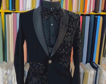 May include: A black tuxedo with a satin lapel and a patterned design on the right side. The tuxedo includes a black shirt, bow tie, and a single-button closure. The background features colorful fabric bolts.