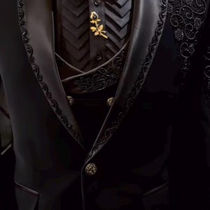 Könnte beinhalten: Ein formeller schwarzer Anzug mit aufwendigen Details. Der Anzug hat eine schwarze Fliege, eine Weste mit dekorativen Knöpfen und eine goldene Rosenbrosche. Die Jacke hat verzierte Stickereien an Revers und Schultern.
