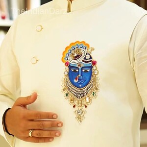 May include: Ivory-colored jacket with a detailed blue face design. The jacket features gold buttons and a decorative emblem with intricate jewel-like accents. The design includes a blue face with a crown and ornate jewelry.