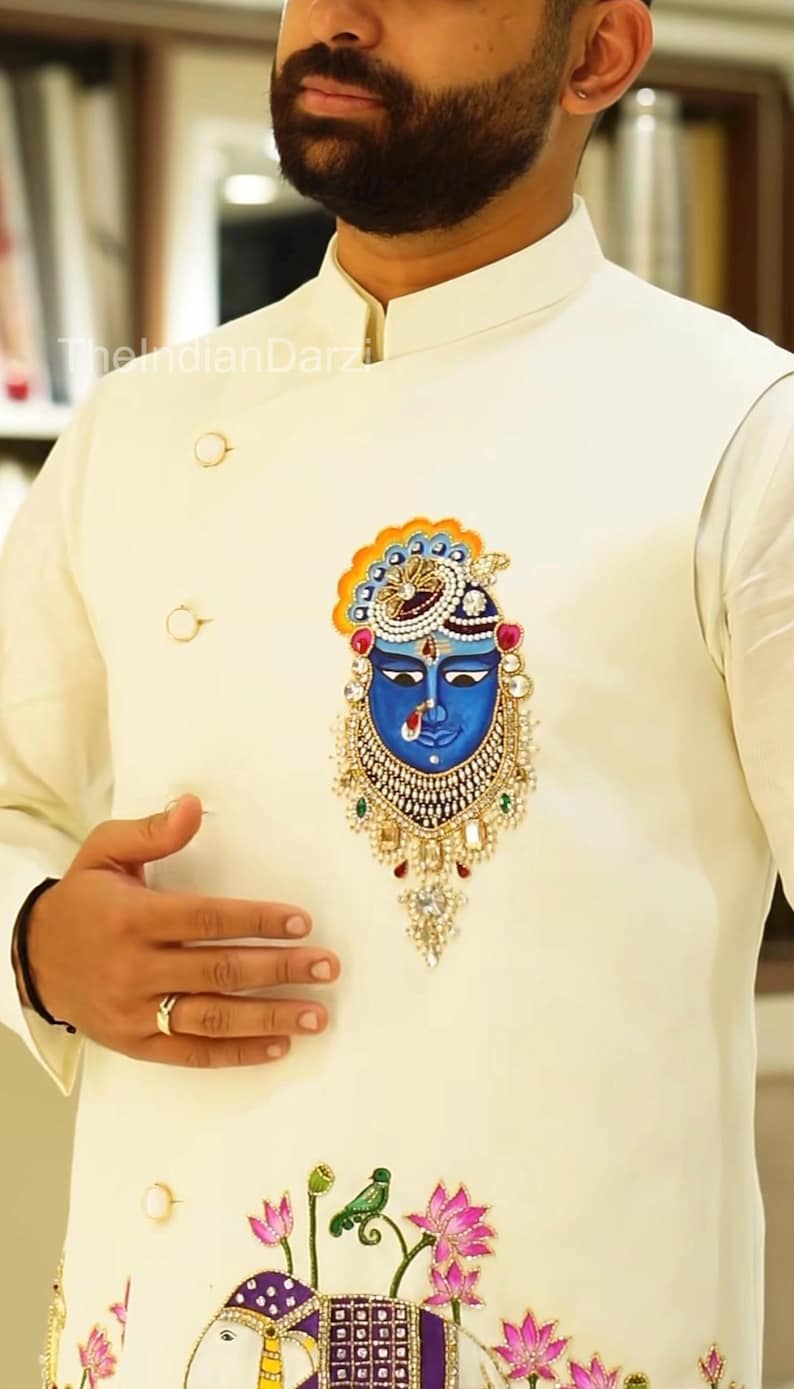 May include: Ivory-colored jacket with a detailed blue face design. The jacket features gold buttons and a decorative emblem with intricate jewel-like accents. The design includes a blue face with a crown and ornate jewelry.