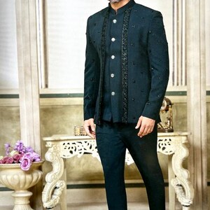 May include: A dark teal suit with a mandarin collar, embellished with black accents. The suit includes a jacket with decorative buttons and matching pants. The person is wearing black dress shoes. The background features a decorative table and floral arrangement.