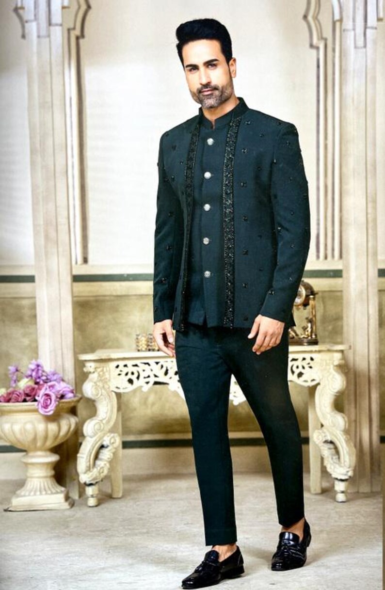 May include: A dark teal suit with a mandarin collar, embellished with black accents. The suit includes a jacket with decorative buttons and matching pants. The person is wearing black dress shoes. The background features a decorative table and floral arrangement.