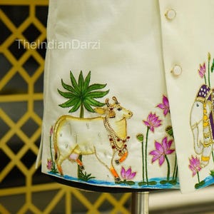 May include: Ivory-colored embroidered jacket with gold and jewel-toned accents. The design features a cow, elephant, lotus flowers, and palm trees. The text "TheIndianDarzi" is visible on the jacket.