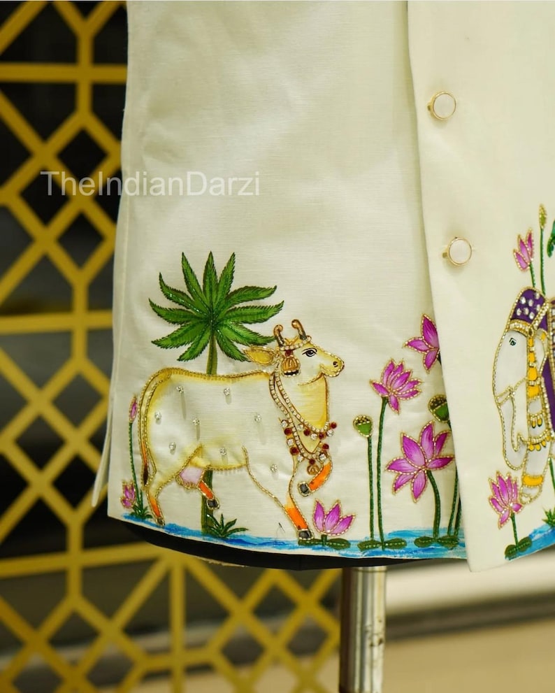 May include: Ivory-colored embroidered jacket with gold and jewel-toned accents. The design features a cow, elephant, lotus flowers, and palm trees. The text "TheIndianDarzi" is visible on the jacket.