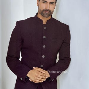 Op de afbeelding: Een donker bordeauxrode blazer met een opstaande kraag en decoratieve knopen. De blazer heeft een subtiel textuurpatroon en een getailleerde pasvorm. De afbeelding bevat de tekst "TheIndianDarzi".